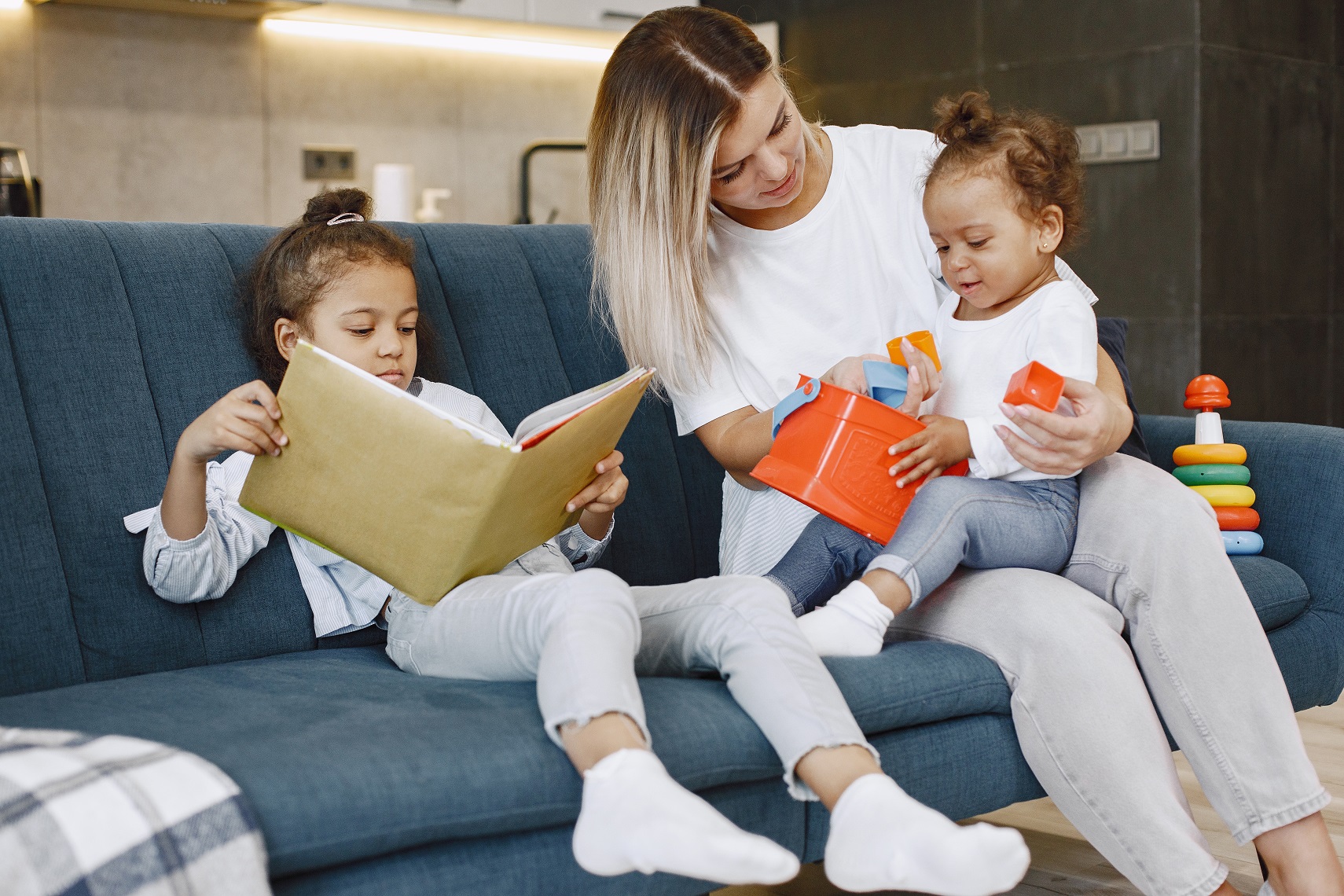Madre e bambini che si rilassano insieme sul divano di casa in soggiorno. Bambine che leggono un libro e giocano a giocattoli.